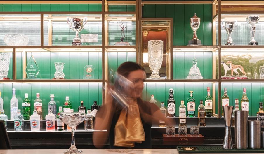 Bartender shaking a cocktail behind a backlit green bar display, with glass trophies on shelving and rows of spirits and glassware arranged neatly in