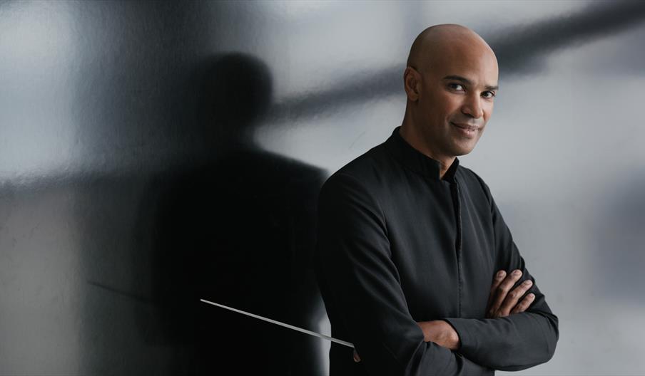 Conductor, Kevin John Edusei leans against a grey wall, and holds his baton while smiling at the camera