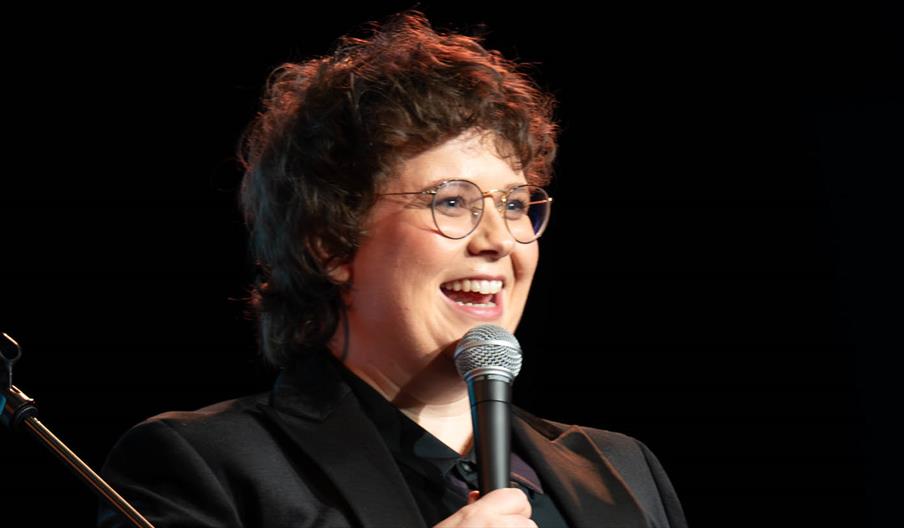 Photograph of a smiling woman, with her mouth slightly open as though in mid laugh. She is holding a microphone and wearing glasses. She has short bro