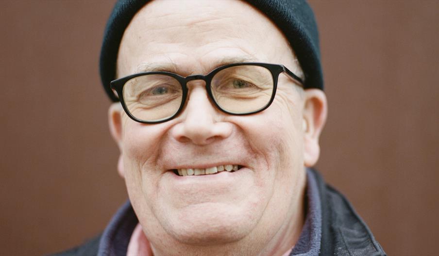 Portrait photograph of an older man wearing a black beanie, black-framed glasses and a dark jacket over a navy fleece and pink shirt. He is smiling at