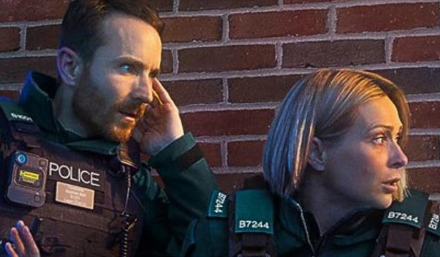 Four police officers in dark green uniforms and tactical vests lean cautiously against a red brick wall at night, looking alert and focused toward the