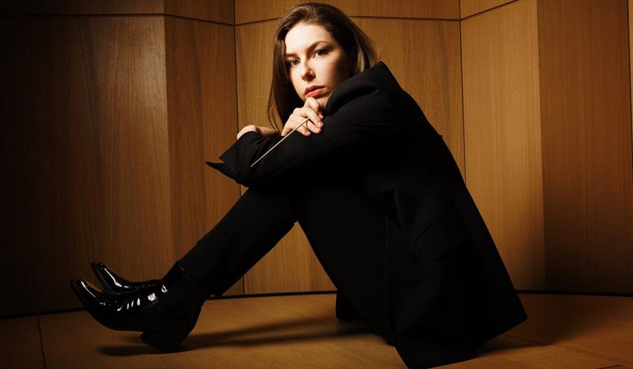 The Ulster Orchestra's new Cheif Conductor sitting on the floor of a concert hall, holding her baton and looking to the camera