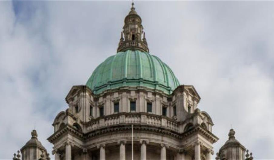 A wide, symmetrical shot shows the grand facade of Belfast City Hall, featuring a large green copper dome, ornate stone columns, and the white marble