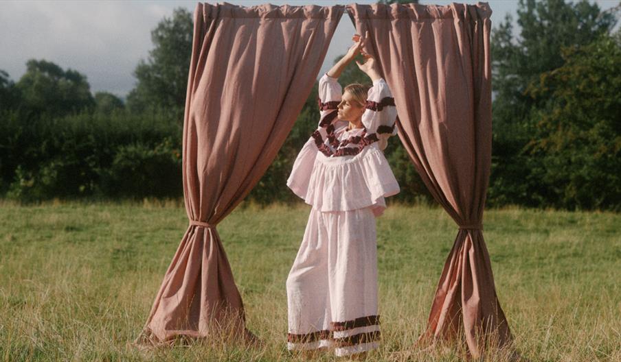 Woman stood with her arms raised above her head in a grass field. She's wearing loose white clothing and is stood in front of a free standing set of c