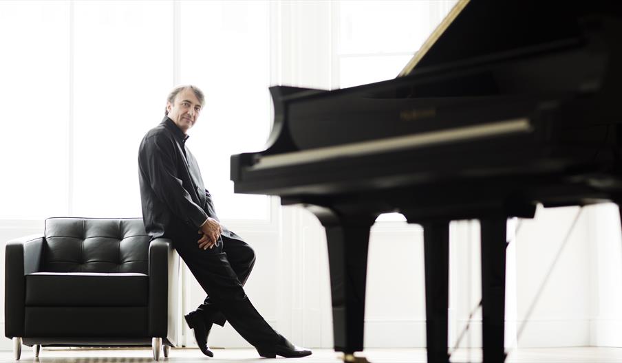 Pianist, Jean-Efflam Bavouzet, leans against a black arm chair with a grand piano in the foreground in a white room