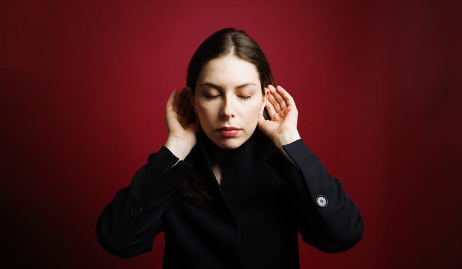 Chief Conductor, Anna Handler stands in black in front of a drawk red background hold up her hands to her ears