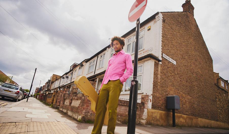 Guitarist, Plínio Fernandes, stands on a street corner in bright pinks and greens holding his guitar in its case