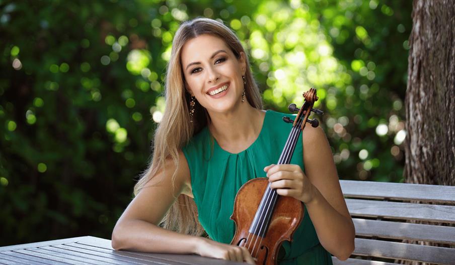 Violinist, Francesca Dego, holds her violin and smiles while sitting outside with trees behind her