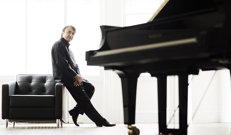 Pianist, Jean-Efflam Bavouzet, leans against a black arm chair with a grand piano in the foreground in a white room