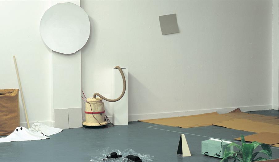 A white room with a grey floor and a collection of objects scattered throughout - (from left to right, top to bottom) a large white paper circle and g