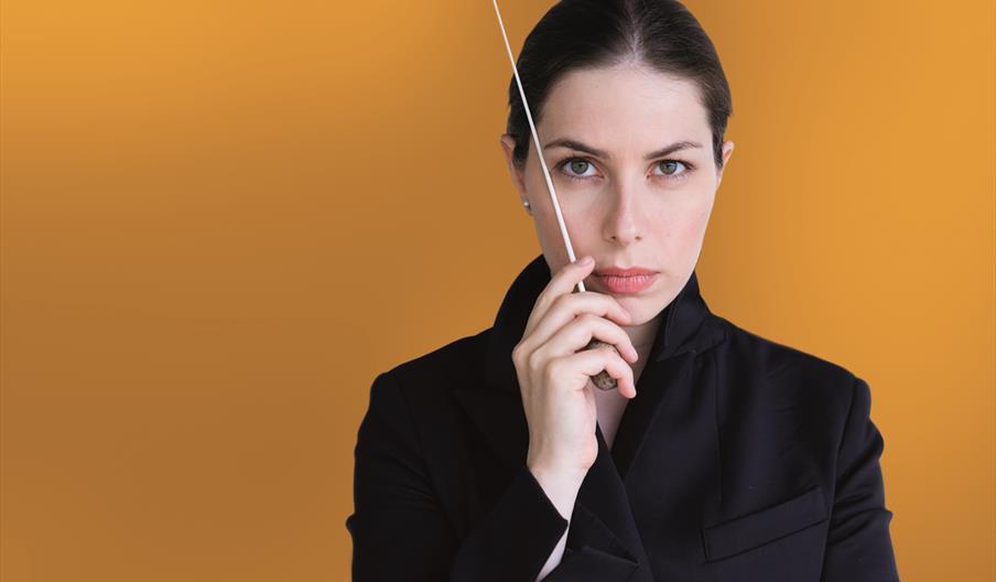 Conductor, Anna Handler stands in front of a yellow background holding her baton and looking into the camera