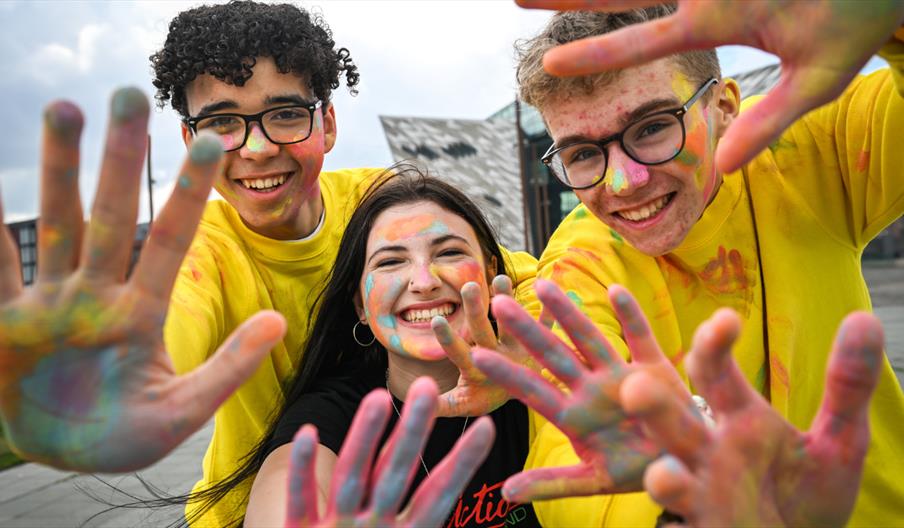 A close up shot of three young people smiling at the camera, their hands messy with colour powder.