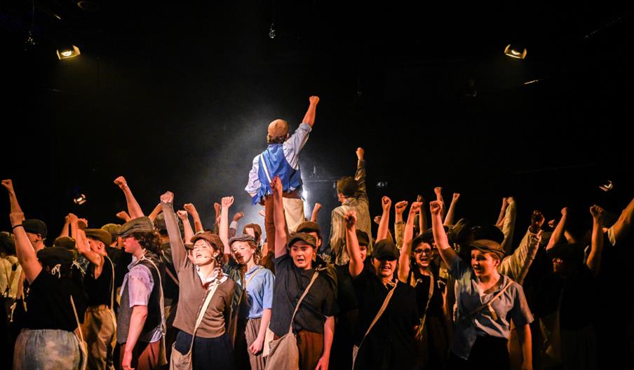 Young people taking part in Disney's 'Newsies'; standing together with their fists in the air, shouting 'Strike!'