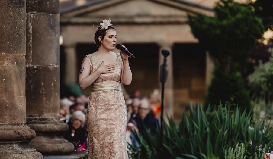 A female singer sings on the South Terrace wearing a gold, patterned dress and floral headwear