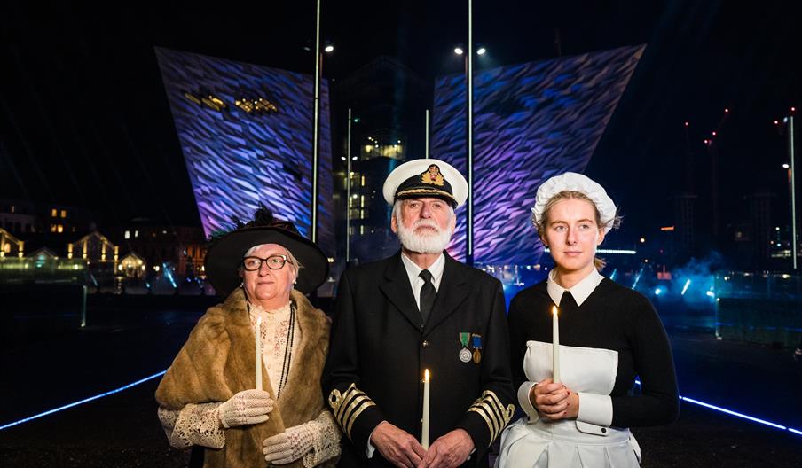 Three people in historical attire stand holding lit candles at night, including a naval officer in uniform flanked by two women in early 20th-century
