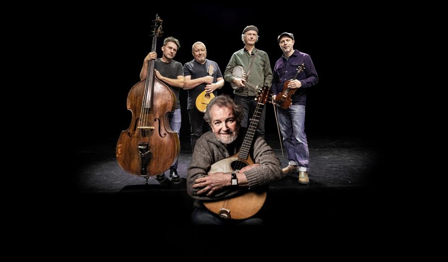 Five musicians pose together on a darkened stage against a black background. One musician sits in the foreground holding an acoustic guitar, while fo