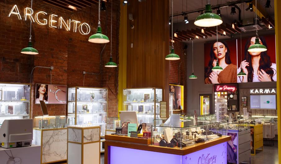 Interior of a modern Argento jewellery store featuring exposed brick walls, warm wood panels and hanging green pendant lights. Illuminated display cas