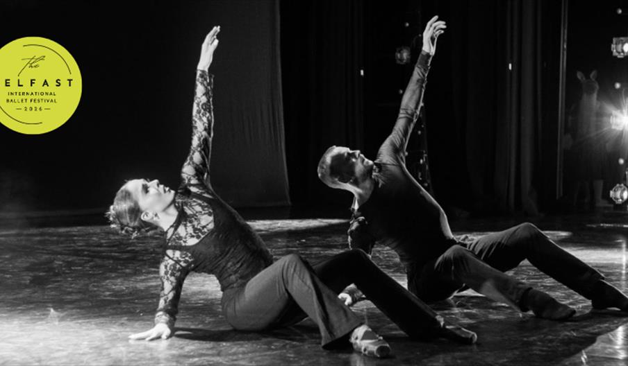 A black and white photograph of a man and a woman performing on stage. They are sitting on the stage floor with their knees bent and their left arms r