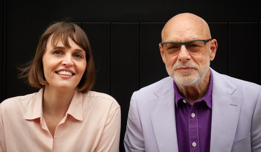 A joint portrait of Bette A and musician Brian Eno sitting side-by-side against a dark paneled background.