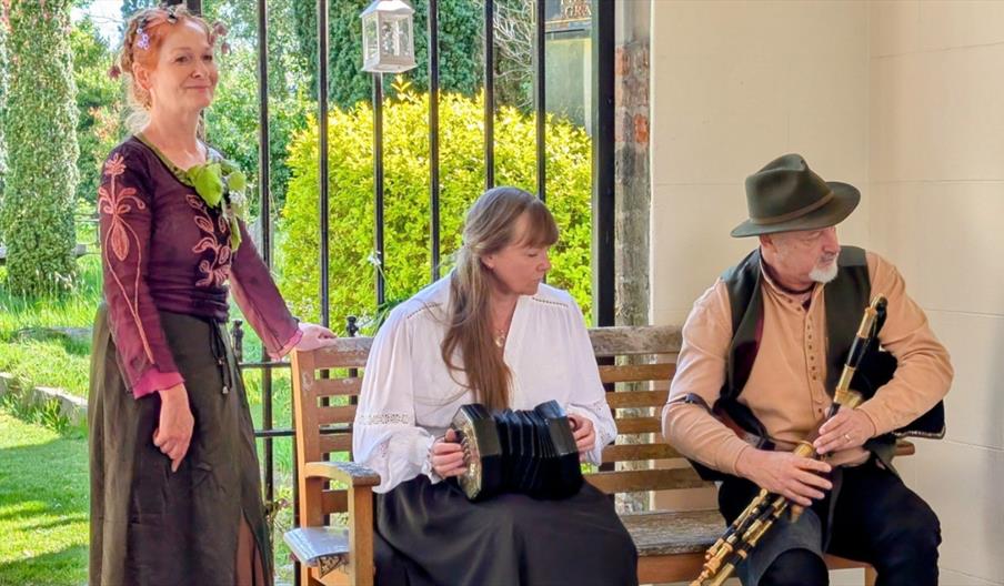 Three people in traditional folk-style clothing sit and stand by a garden bench, with one holding a concertina and another holding bagpipes.
