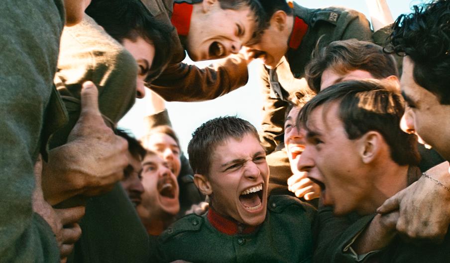 A jubilant group of young men in dark uniforms crowd together, shouting and laughing in a celebratory huddle beneath a bright sky.