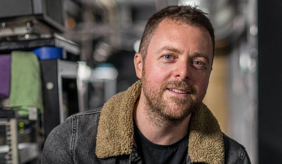 A bearded man in a black jacket with a tan fleece collar smiles slightly at the camera in a busy workshop, conveying a warm, approachable mood.