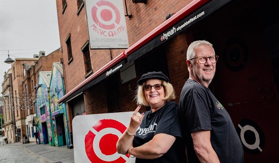 Two smiling adults standing back-to-back outside Oh Yeah Music Centre on a cobblestone street,