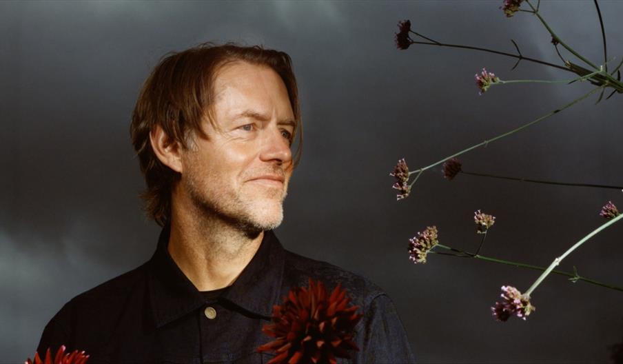A profile portrait of musician Ed O'Brien looking thoughtfully to the right, surrounded by dark red dahlias and tall purple verbena against a dramatic