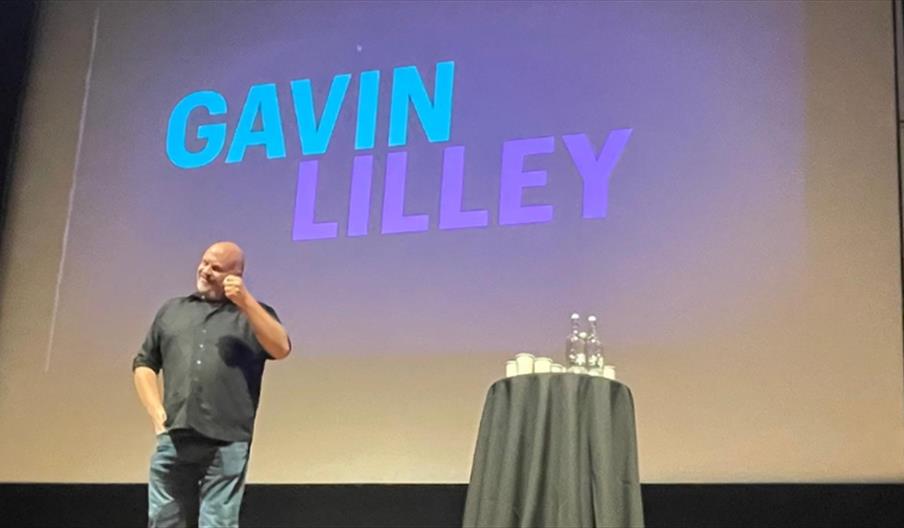 Photo of Gavin Lilley on stage. His name is on a stage behind him