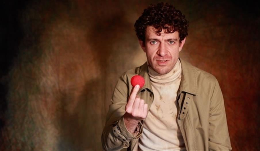 Photo of Sam Blythe looking at us. He is holding a red nose.