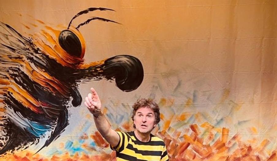 Colin, the main actor is dressed in black and yellow striped clothes. He is pointing at something in the sky. Behind him,  the set displays a large wh