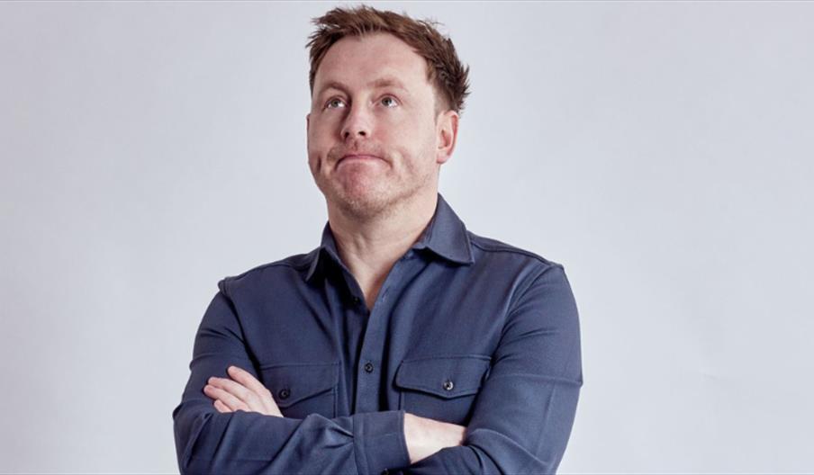 A youngish man with short ginger hair standing against a grey background with his arms crossed and a fed up expression on his face.