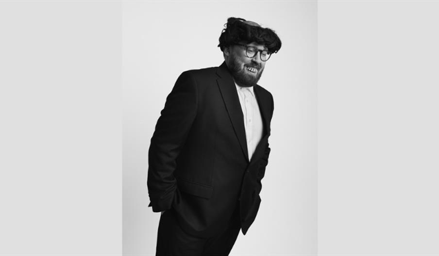 Black and white picture of John Kearns hands in his pockets. He is wearing a wig and fake teeth