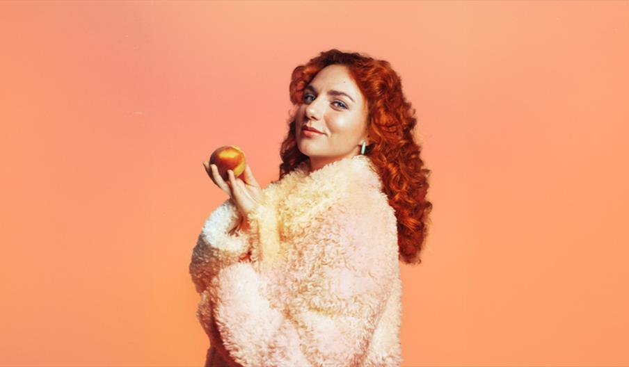 Ania Magliano is looking at us holding a peach in front of a peach-coloured background. She has a furry peach coat