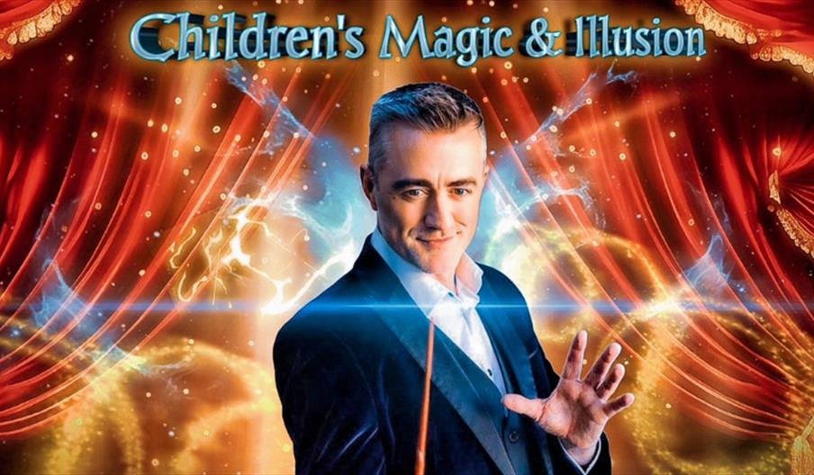 Photo of Joe the magician smiling and holding his magic wand. The background is the red theatre curtains with a gold magic trail.