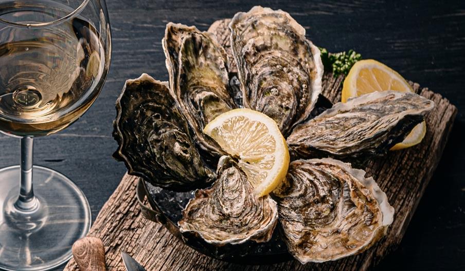 A platter of fresh oysters with slice of lemon on a wooden board next to a glass of white wine