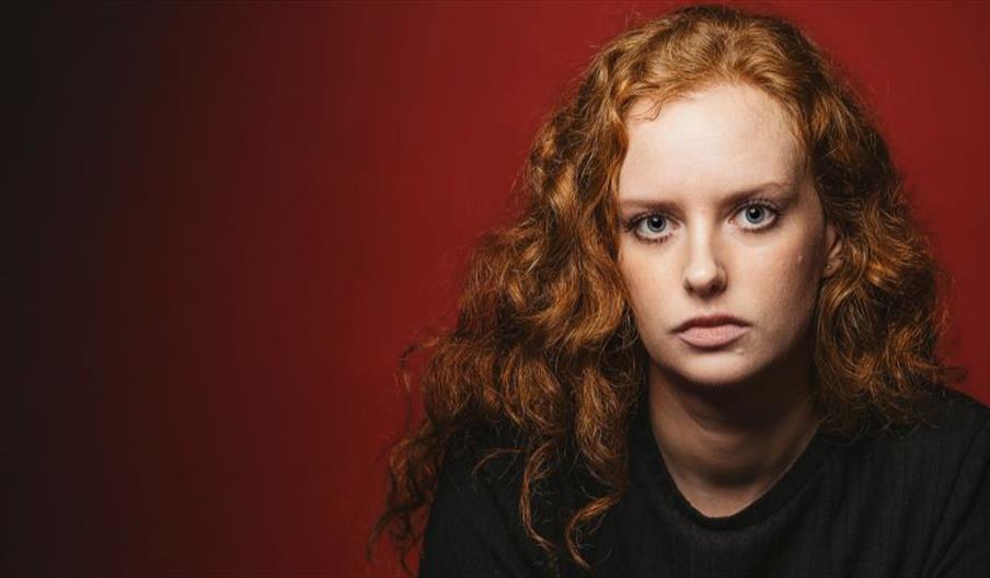 A young woman with curly ginger hair and blue-grey eyes looks with a serious gaze right at the viewer. Dark red background which ombres into black to