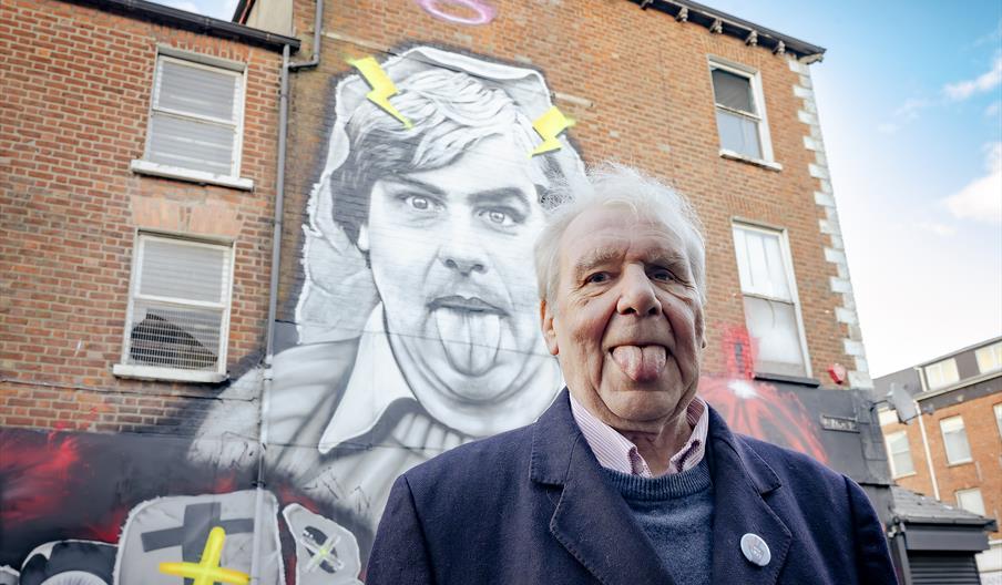 Picture shows Terri Hooley standing in front of a mural depicting himself with his tongue out