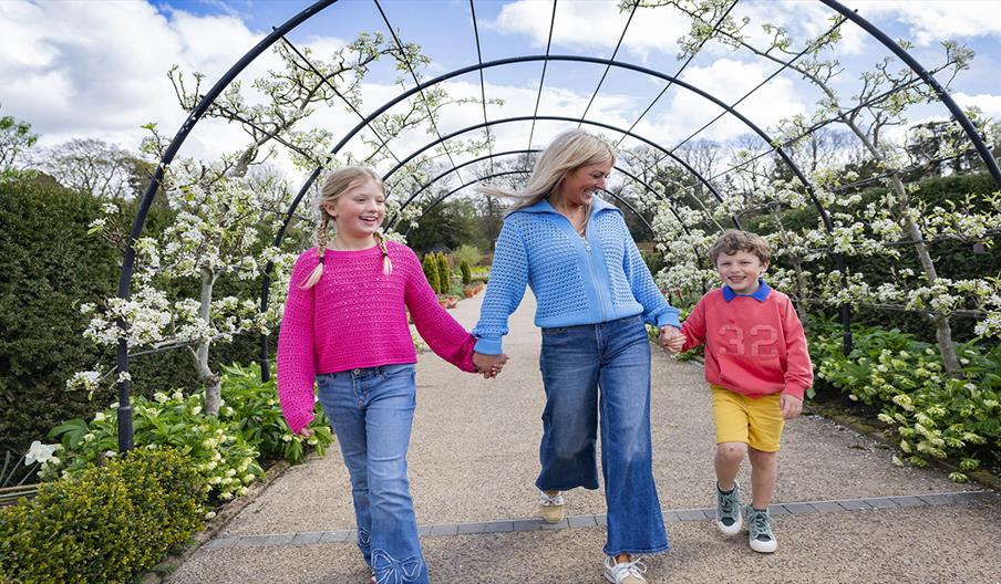 mother and two children enjoying a lovely spring day at Hillsborough Castle and Gardens, surrounded by blossoms and trees under a partly cloudy blue s