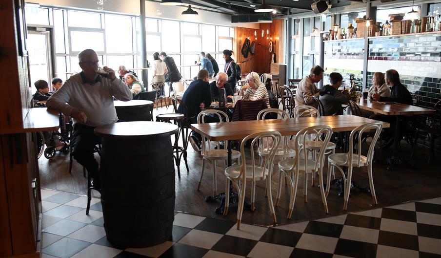 Groups of people sat at various tables within a restaurant, there are windows on all the walls and a black and white checker floor.