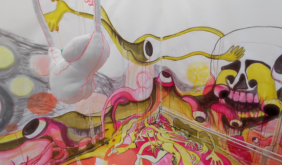 a corner of a room with walls and floor covered in colourful, wavy illustrative designs resembling misshaped body parts with a textile 3d object hangi