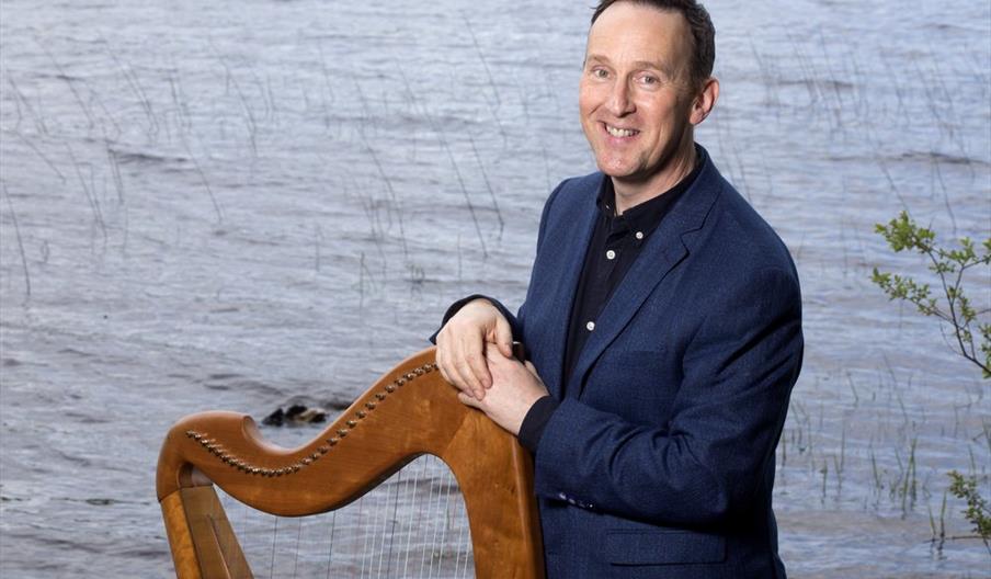 Michael Rooney standing with a Harp against a backdrop of water