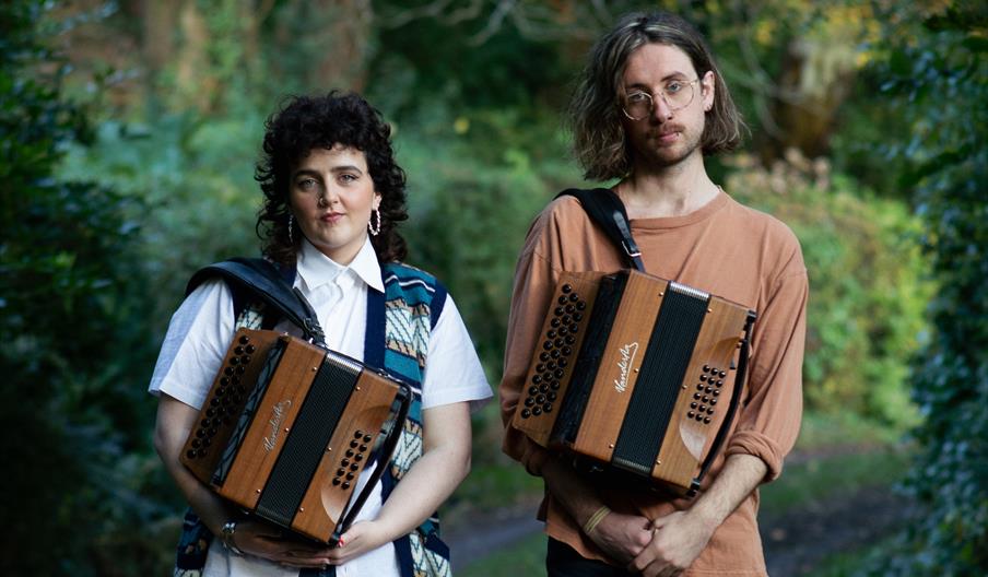 Moya & Archie standing against a backdrop of trees and greenery, both holding their accordions