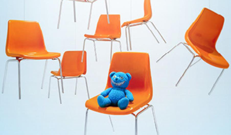 Several orange chairs floating in the air. The chair to the fore of the image has a ragged blue teddy on it.