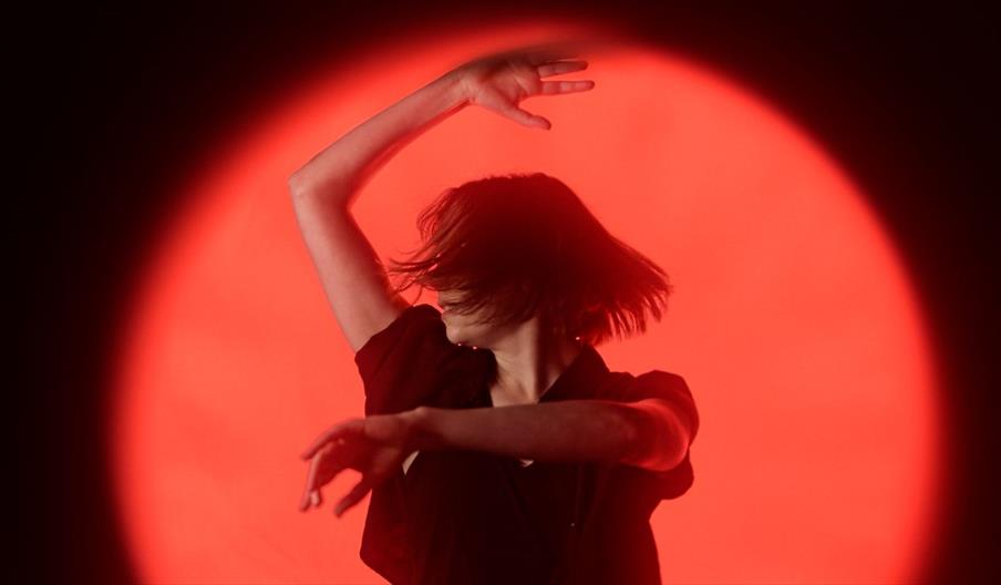 A dancer with short hair captured mid-movement in silhouette, arms raised gracefully against a vivid red circular spotlight.
