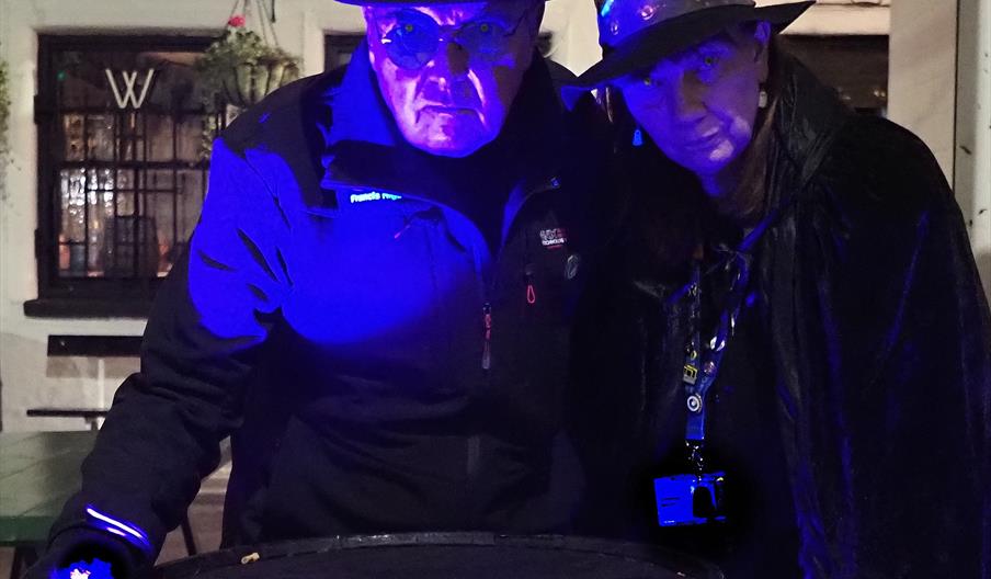 Two people wearing dark coats and black hats lean toward a large round barrel in an outdoor nighttime setting. Blue and purple lighting casts a dramat