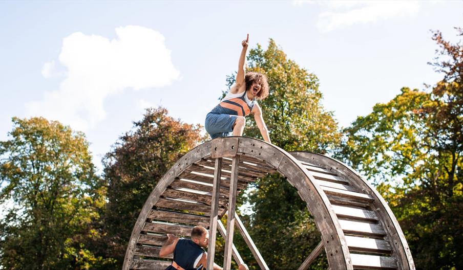 Outdoor performance showing a man with curly hair perched atop a large wooden hamster-wheel structure pointing joyfully into the air while another per