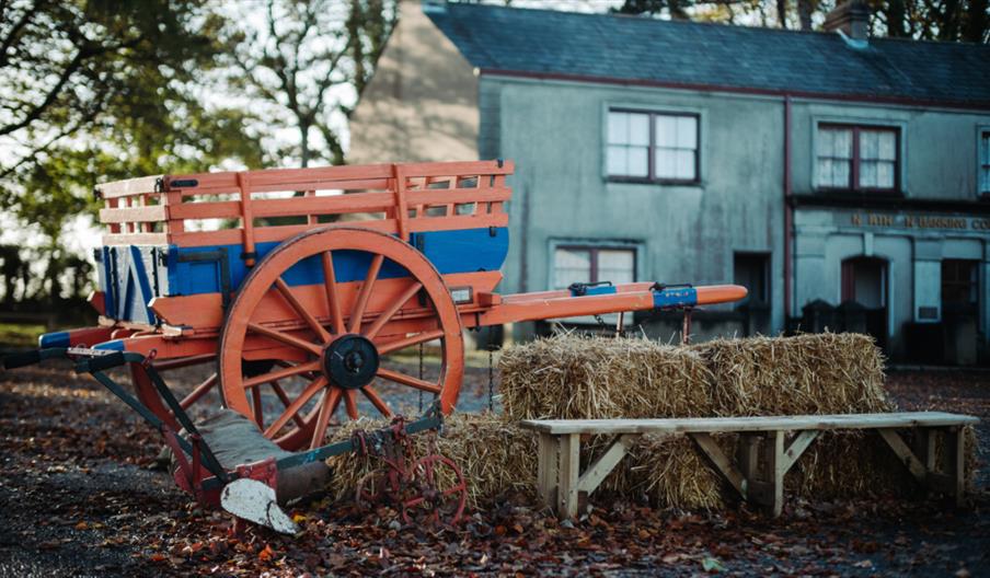 A blue and red wagon leaning on hay barrels with grey houses and trees in the background situated in the diamond at ulster folk museum