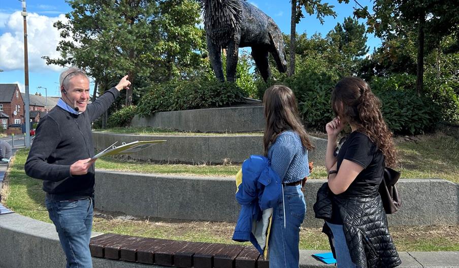 Tour guide pointing to steel sculpture of lion with 2 people looking