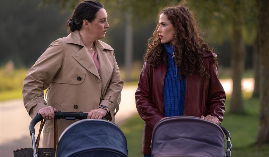 Two women pause beside baby strollers on a tree-lined path, facing each other in a serious conversation; the mood feels tense and concerned.